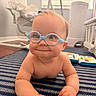 baby, child, glasses, blue_glasses, smile, grinning, striped_rug, nursery, toy, bracelet, floor, indoor, infant, crawling, cute, playtime, furniture, window_blinds, soft_lighting, happy
