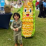 Anabia is registered to the contest to win money with this photo: child, smiling, curly_hair, green_jumpsuit, brown_shoes, corn_costume, mascot, grass, outdoor, event, people, booth, tent, happy, costume, fun, daytime, park, celebration, portrait