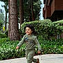 child, boy, running, smiling, hoodie, pants, brown_shoes, curly_hair, outdoor, sidewalk, trees, bushes, greenery, building, daytime, playful, happy, casual_clothing, nature, urban