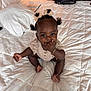 Layla is registered to the contest to win money with this photo: baby, child, infant, pigtails, bow, bed, white_bedspread, pillow, bracelet, hand, foot, sitting, cute, indoors, room, furniture, curious, playful, person, portrait