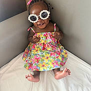 Layla is registered to the contest to win money with this photo: baby, child, girl, sunglasses, flower_bows, floral_dress, smile, teeth, feet, bed, corner, white_sheet, skin, happy, cute, indoors, portrait, fashion, accessories, hair