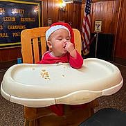 Mlijah joined the competition — help win amazing prizes! toddler, child, high_chair, santa_hat, red_clothing, wooden_chair, crumbs, indoor, wood_paneling, american_flag, banner, baseball, holiday, festive, cute, eating, person, face, baby, cozy