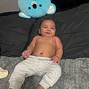 Antonio is registered to the contest to win money with this photo: baby, infant, smiling, bare_chest, gray_blanket, black_pillow, plush_toy, blue_koala, soft_texture, cute, relaxed, indoors, child, person, sleepwear, foot, hand, cozy, comfortable, happy