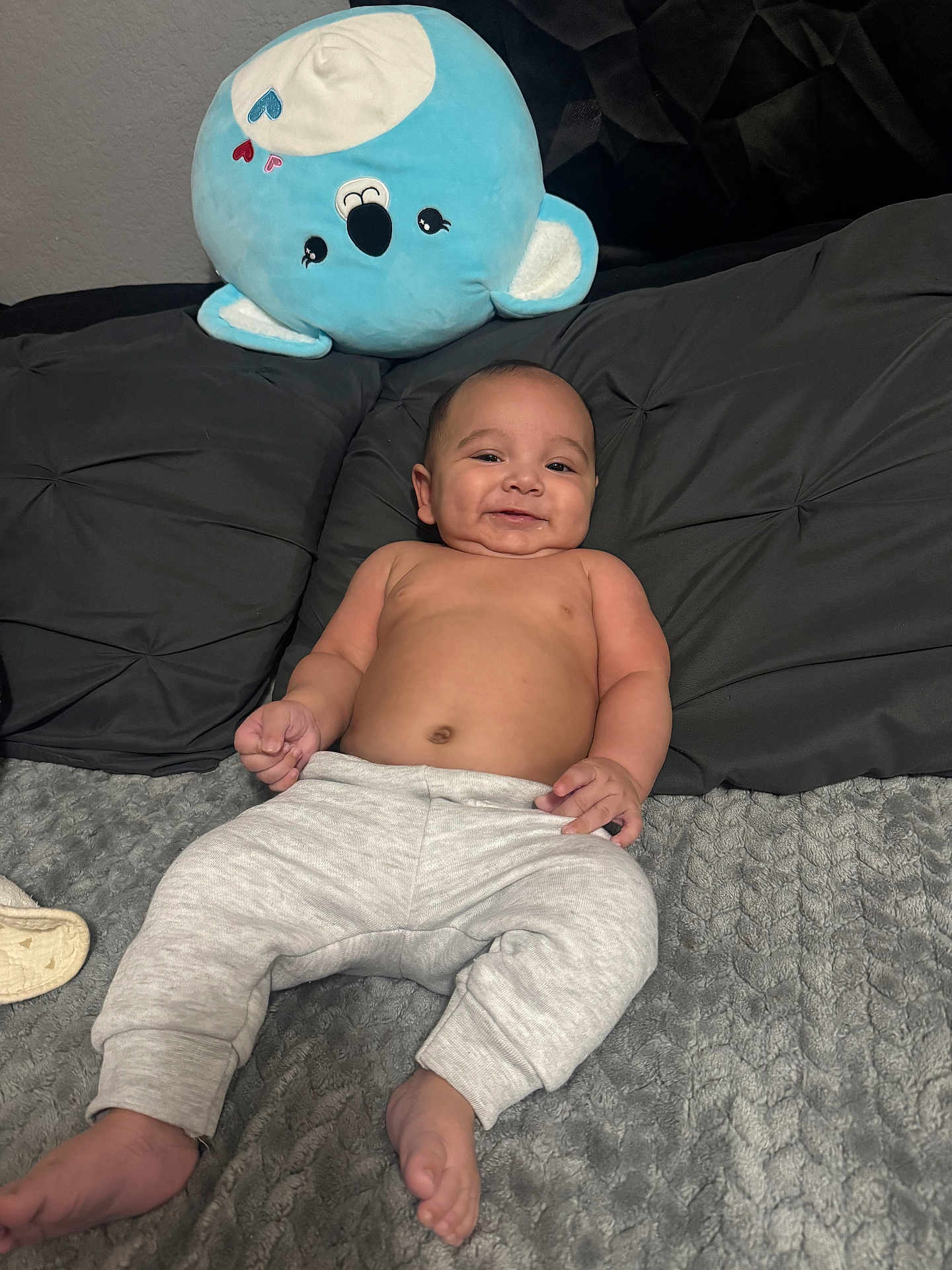 Antonio is registered to the contest to win money with this photo: baby, infant, smiling, bare_chest, gray_blanket, black_pillow, plush_toy, blue_koala, soft_texture, cute, relaxed, indoors, child, person, sleepwear, foot, hand, cozy, comfortable, happy