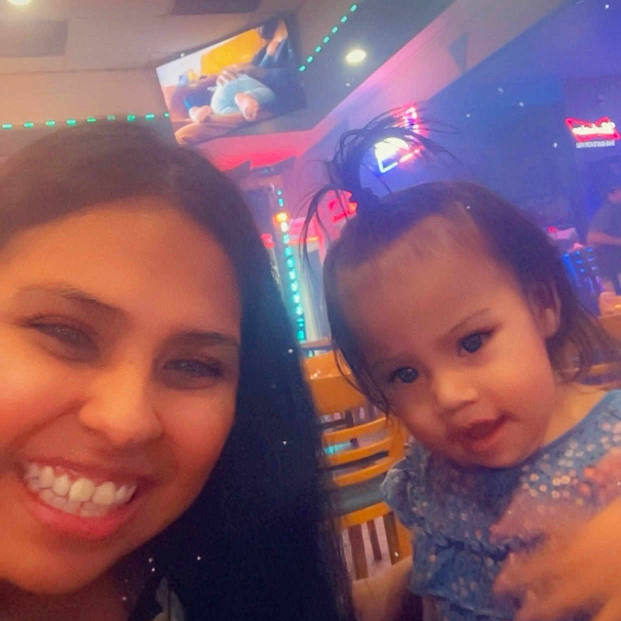April joined the competition — help win amazing prizes! woman, toddler, child, selfie, portrait, smiling, holding_child, indoor, restaurant, neon_lights, colorful_lighting, blue_dress, sparkly_clothing, long_hair, baby, face, family, mother, background_lights, casual_photo
