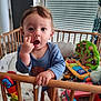 Lyam a rejoint le concours — aidez-le/la à gagner de superbes lots ! baby, child, crib, toy, indoor, onesie, blue_clothing, wooden_crib, playroom, curious, standing, furniture, window, blinds, colorful, expression, hand, face, person, toddler
