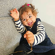 Azalia a rejoint le concours — aidez-le/la à gagner de superbes lots ! toddler, child, smile, curly_hair, glasses, striped_shirt, black_tights, blue_dress, couch, indoor, happy, playful, person, face, hand, sitting, cute, young_child, home, fun