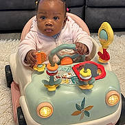 Ayla is registered to the contest to win money with this photo: baby, toy_car, indoor, child, cute, play, steering_wheel, hair_ties, pink_jacket, car_toy, floor_rug, couch, child_playing, baby_toy, infant, colorful, hands, expression, seat, front_view