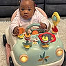baby, toy_car, indoor, child, cute, play, steering_wheel, hair_ties, pink_jacket, car_toy, floor_rug, couch, child_playing, baby_toy, infant, colorful, hands, expression, seat, front_view