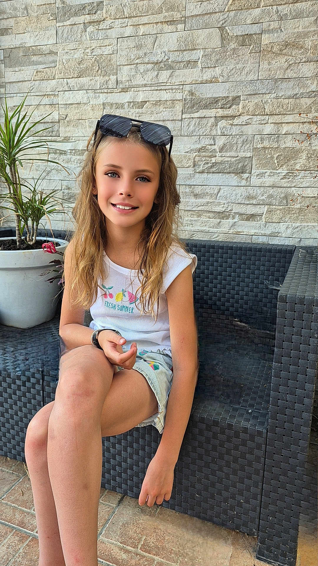 Kahina participe au concours pour gagner de l'argent avec cette photo : child, girl, smiling, sunglasses, white_tank_top, shorts, long_hair, sitting, outdoor_furniture, wicker_couch, plant, potted_plant, stone_wall, tile_floor, casual, sunny, portrait, young, happy, relaxed