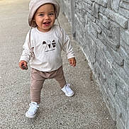 Liam joined the competition — help win amazing prizes! toddler, child, hat, bucket_hat, smiling, walking, sidewalk, stone_wall, outdoor, casual_clothing, white_shoes, pants, long_sleeve_shirt, holding_object, rock, young_child, happy, person, daylight, concrete