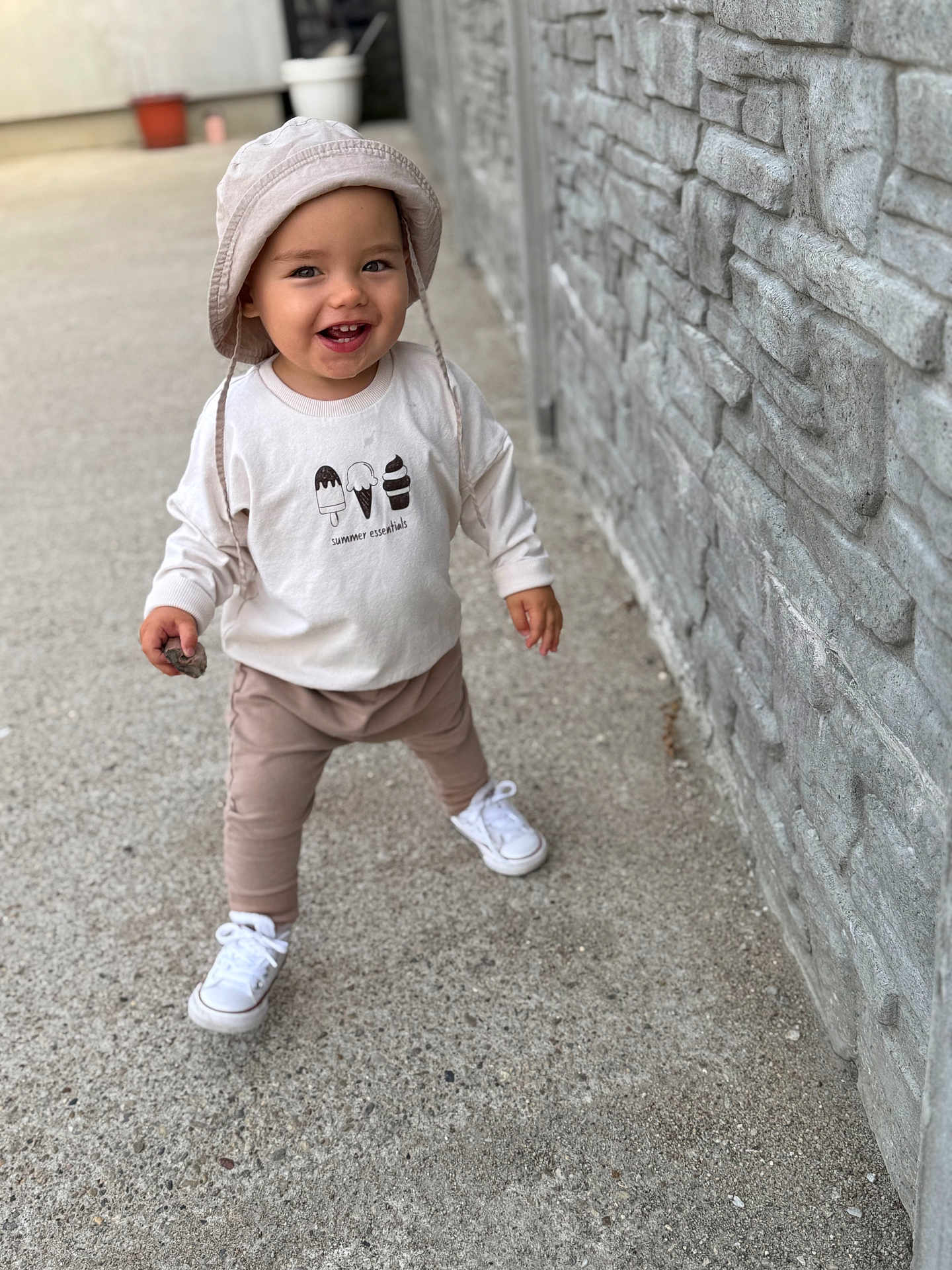 Liam joined the competition — help win amazing prizes! toddler, child, hat, bucket_hat, smiling, walking, sidewalk, stone_wall, outdoor, casual_clothing, white_shoes, pants, long_sleeve_shirt, holding_object, rock, young_child, happy, person, daylight, concrete