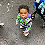 blanket, blue_bag, bubbles, buzz_lightyear, casual_clothing, child, curious, curly_hair, cute, hoodie, looking_up, outdoor, person, playful, sidewalk, small_child, stroller, toddler, wet_pavement, white_shoes