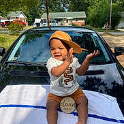 Kyyeer is registered to the contest to win money with this photo: baby, child, toddler, hat, cap, clapping, smiling, outdoor, car, black_car, towel, milestone_sign, eleven_months, clothing, shorts, white_shirt, sunny, suburban, trees, house