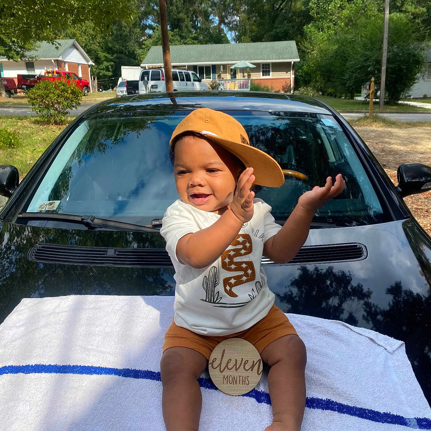 Kyyeer is registered to the contest to win money with this photo: baby, black_car, cap, car, child, clapping, clothing, eleven_months, hat, house, milestone_sign, outdoor, shorts, smiling, suburban, sunny, toddler, towel, trees, white_shirt