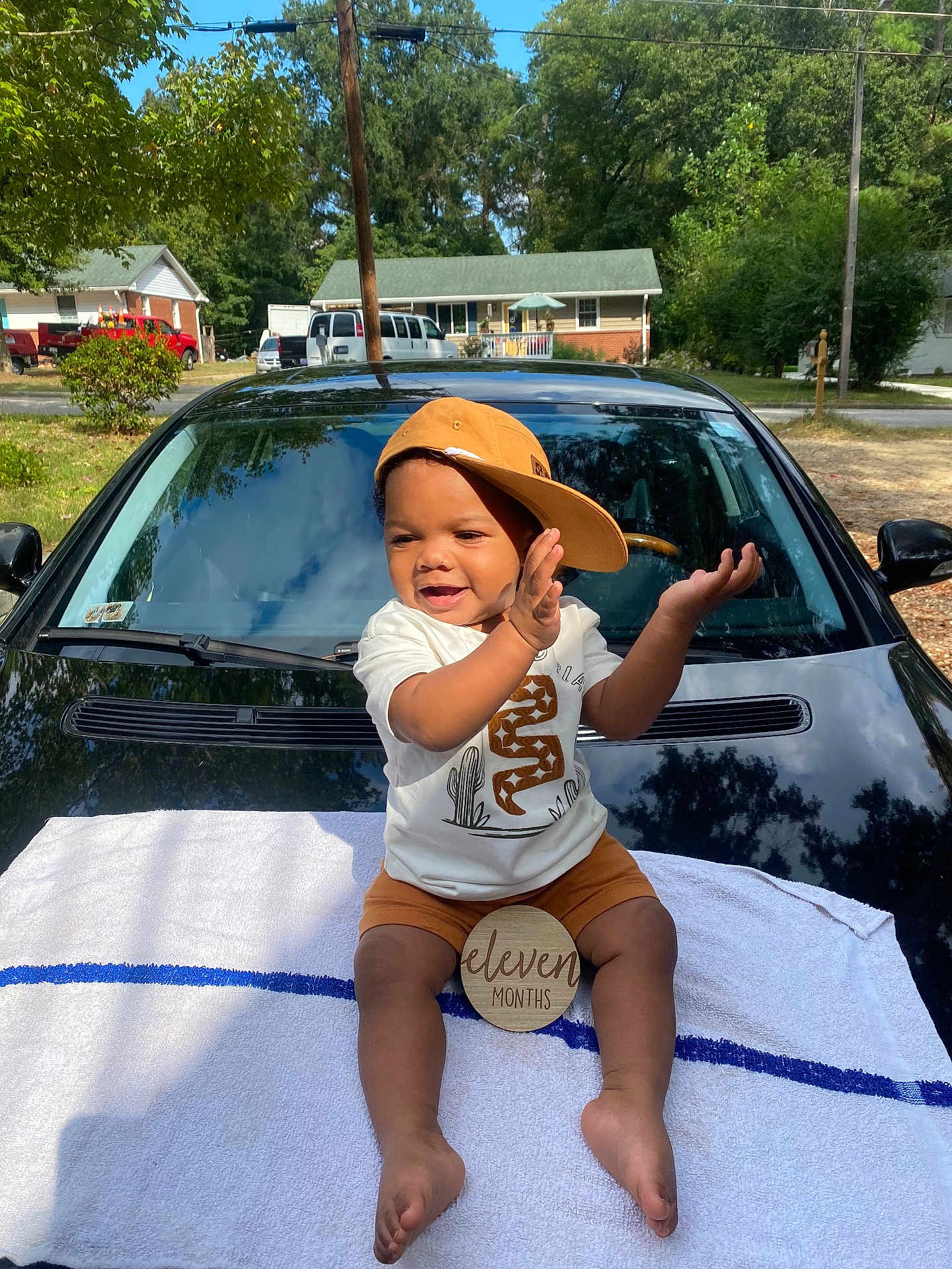 Kyyeer is registered to the contest to win money with this photo: baby, child, toddler, hat, cap, clapping, smiling, outdoor, car, black_car, towel, milestone_sign, eleven_months, clothing, shorts, white_shirt, sunny, suburban, trees, house