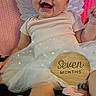baby, infant, child, smiling, happy, angel_costume, wings, headband, flower, tutu, dress, milestone, seven_months, portrait, lying_down, indoor, pink_blanket, wooden_sign, bare_feet, celebration