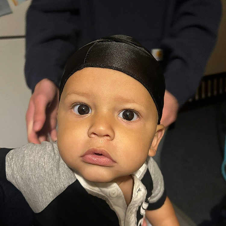 Josiah is registered to the contest to win money with this photo: adult_hand, baby, black_cap, child, close_up, clothing, curious, expression, eyes, face, hand, human, indoor, person, portrait, shirt, skin, support, toddler, young_child