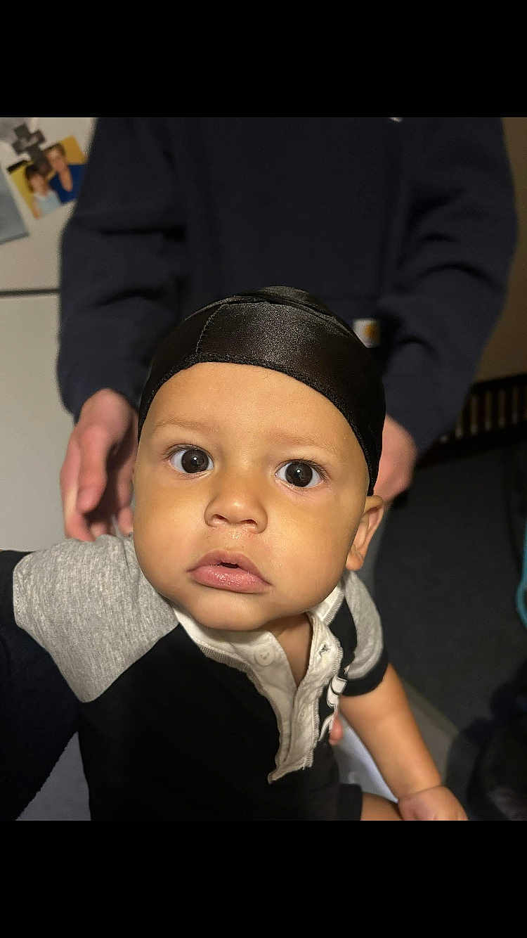 Josiah is registered to the contest to win money with this photo: toddler, child, face, eyes, black_cap, shirt, indoor, person, hand, adult_hand, curious, close_up, portrait, baby, skin, expression, clothing, young_child, human, support