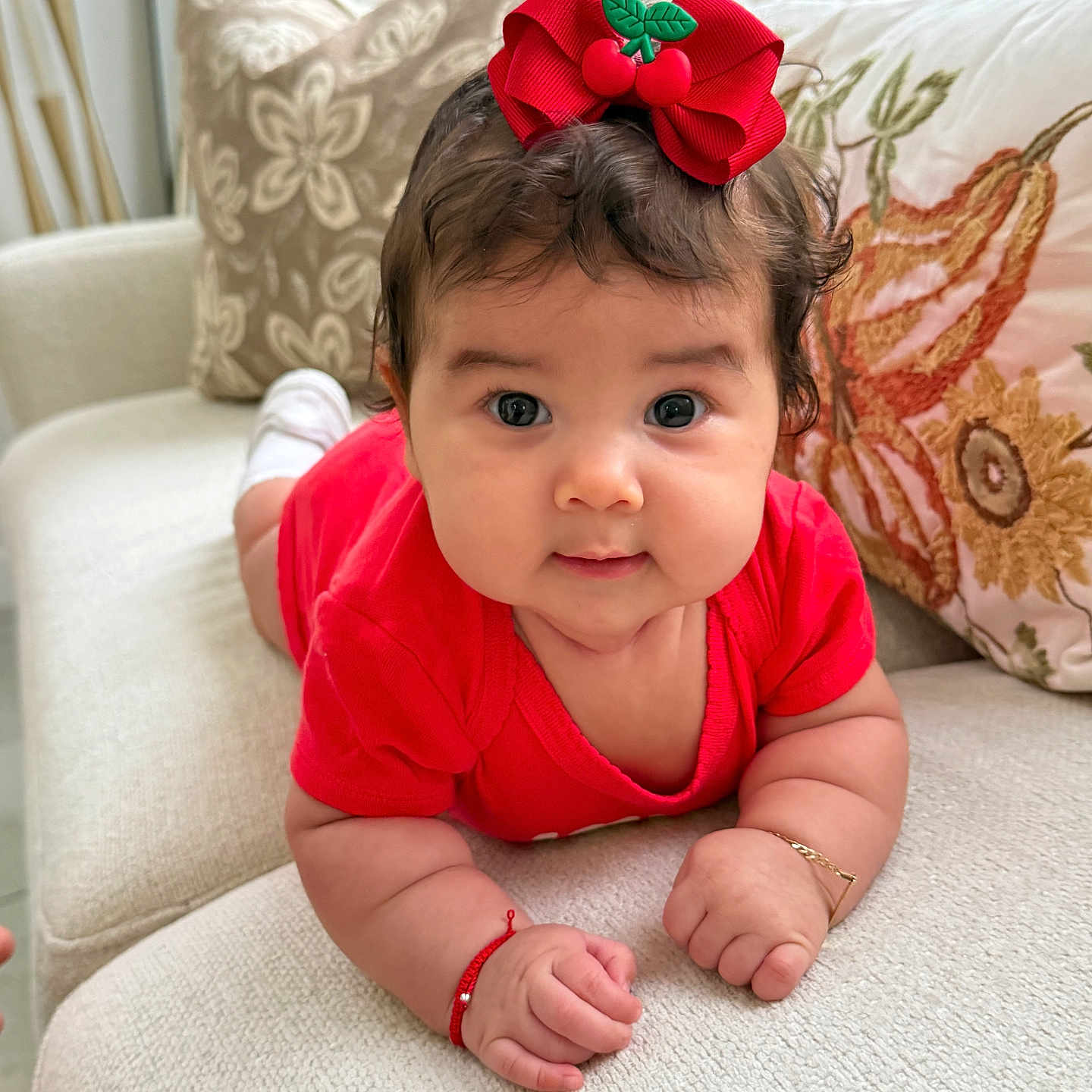 Reina Antonella is registered to the contest to win money with this photo: baby, child, infant, portrait, smiling, wide_eyes, red_outfit, hair_bow, cherry_bow, sitting, crawling, sofa, couch, cushions, indoor, home, close_up, adorable, bracelet, photography