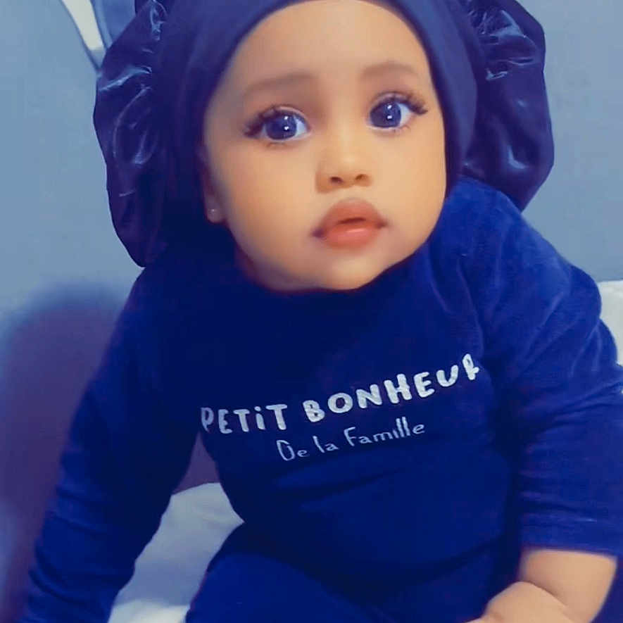 Aisha Saw participe au concours pour gagner de l'argent avec cette photo : baby, bed, blue_clothing, bonnet, child, cozy, curious, cute, expression, face, hand, indoors, infant, person, pillow, portrait, sitting, soft_lighting, text_on_clothing, young_child