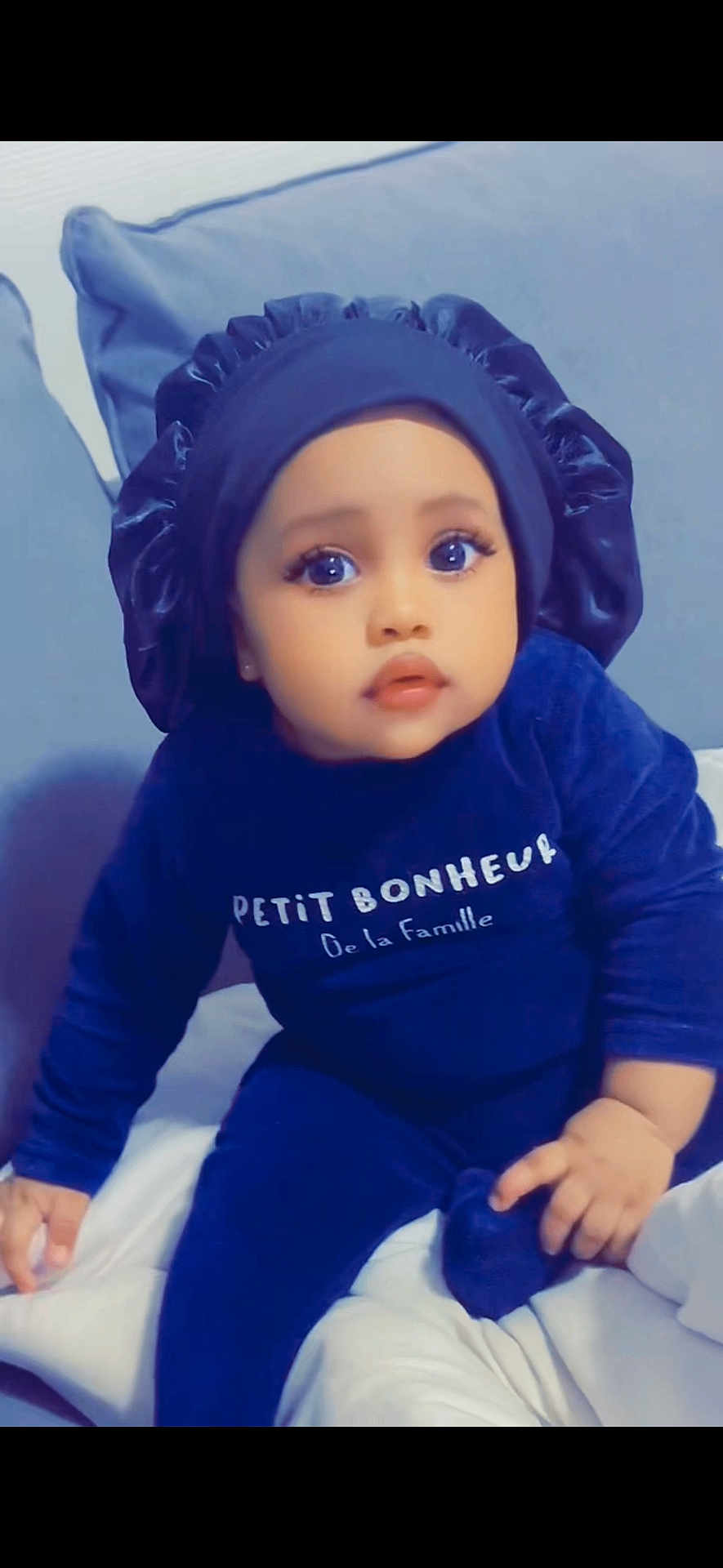 Aisha Saw participe au concours pour gagner de l'argent avec cette photo : baby, child, infant, blue_clothing, bonnet, bed, pillow, cute, portrait, indoors, person, sitting, expression, face, hand, young_child, cozy, soft_lighting, text_on_clothing, curious