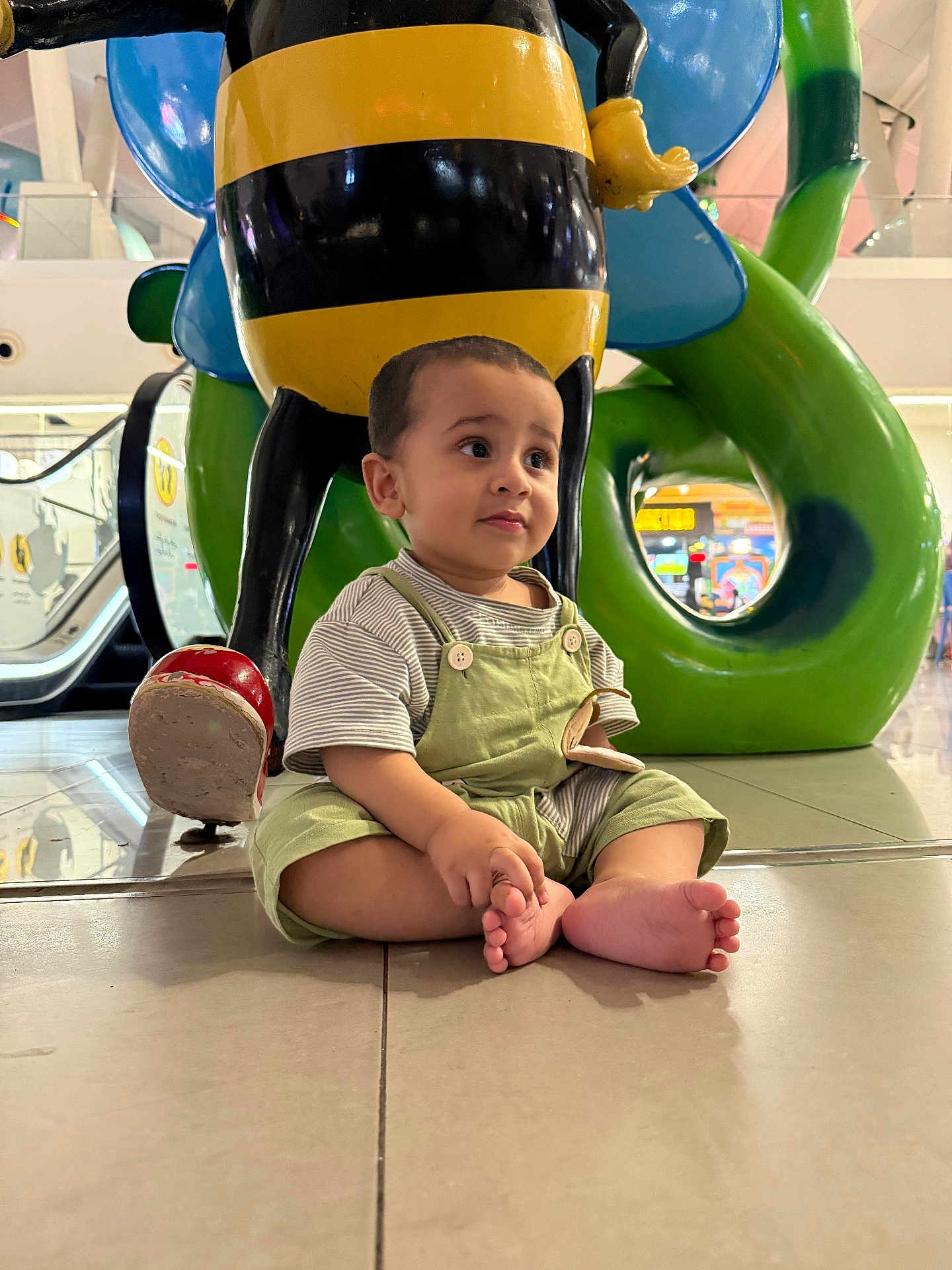 Muhammad joined the competition — help win amazing prizes! toddler, child, barefoot, overalls, striped_shirt, sitting, indoor, statue, bee_statue, colorful, green, yellow, red, floor, tiles, mall, playful, curious, young_child, sculpture