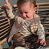 child, toddler, baby, sitting, chair, woven_chair, portrait, outdoor, sunlight, shadows, smiling, happy, face, hair, clothing, vest, pants, sneakers, raised_hand, casual
