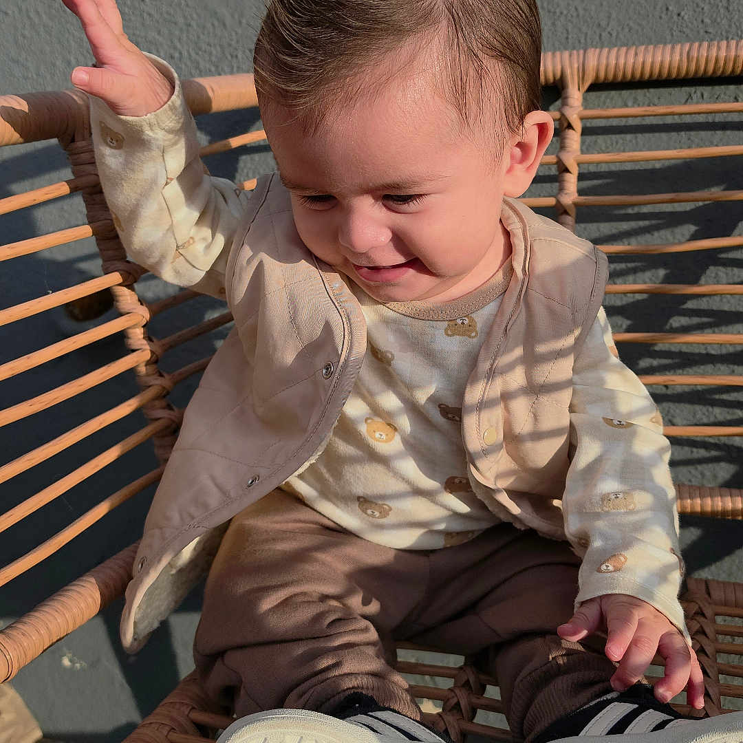 Liam Santos joined the competition — help win amazing prizes! child, toddler, baby, sitting, chair, woven_chair, portrait, outdoor, sunlight, shadows, smiling, happy, face, hair, clothing, vest, pants, sneakers, raised_hand, casual