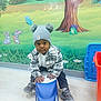 toddler, child, hat, pom_poms, toy_car, indoor, floor, wall_mural, cartoon_animals, rabbit, butterfly, frog, tree, grass, flowers, sneakers, jacket, pants, playtime, smile