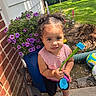 Safia joined the competition — help win amazing prizes! toddler, child, outdoor, garden, flowers, purple_flowers, flower_pot, watering_can, toy, pigtails, pink_shirt, purple_shoes, green_grass, sidewalk, brick_wall, sunlight, playful, curious, person