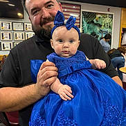 Laken is registered to the contest to win money with this photo: baby, background, beard, blue_dress, certificate, chair, child, clothing, decor, event, face, headband, holding, indoor, man, people, person, portrait, smile, table
