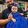 baby, background, beard, blue_dress, certificate, chair, child, clothing, decor, event, face, headband, holding, indoor, man, people, person, portrait, smile, table