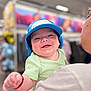 Ariel joined the competition — help win amazing prizes! baby, child, smiling, hat, blue_hat, green_shirt, person, adult, holding, indoor, store, shopping, blurred_background, portrait, happy, cute, infant, closeup, face, apparel