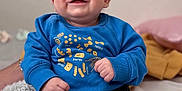Ariel is registered to the contest to win money with this photo: baby, smiling, blue_sweater, pasta_illustration, sitting, bed, blanket, pillow, happy, infant, clothing, person, indoors, cute, portrait, child, soft_toys, cozy, casual, relaxed