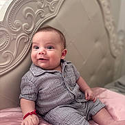 Ariel is registered to the contest to win money with this photo: baby, infant, striped_clothing, bed, pink_bedspread, headboard, ornate_furniture, carving, bracelet, happy, curious, portrait, indoor, child, person, sitting, closeup, cute, smile, soft_lighting