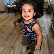 Izmael is registered to the contest to win money with this photo: toddler, child, indoor, wooden_floor, dinosaur_shirt, curly_hair, smiling, playful, person, socks, furniture, chair, lamp, window, home, flooring, boy, happy, cute, casual
