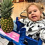 child, toddler, shopping_cart, pineapple, mickey_mouse, sweater, blonde_hair, pigtails, mall, floor_tiles, blue_cart, smiling, person, indoor, retail, hand, bag, people, casual_clothing, shopping
