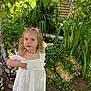 child, girl, white_dress, nature, greenery, plants, outdoor, tree, leaves, sunlight, necklace, portrait, young_child, grass, shoes, daylight, curious, blonde_hair, standing, garden