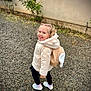 child, smiling, outdoor, gravel, backpack, jacket, footwear, happy, walking, building, wall, window, plant, nature, casual_clothing, young_child, blonde_hair, playful, small, cute