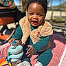 baby, child, smiling, outdoor, toy, blanket, vest, knitwear, happy, sitting, face, person, footwear, sunlight, play, infant, cute, colorful, warm_clothing, portrait