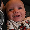 Xavier is registered to the contest to win money with this photo: baby, infant, face, expression, smile, onesie, clothing, pattern, hand, person, closeup, cute, child, young, indoor, dark_background, portrait, happy, skin, eyes