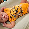 Xavier is registered to the contest to win money with this photo: baby, infant, onesie, orange_clothing, cartoon_print, crib, mattress, wooden_crib, child, face, portrait, gaze, lying_down, indoors, cute, young_child, person, skin, head, smile