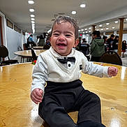 Augustine joined the competition — help win amazing prizes! baby, toddler, child, smiling, bow_tie, formal_attire, white_shirt, black_pants, socks, wooden_table, indoor, party, banquet_hall, people, chairs, ceiling_lights, happy, portrait, sitting, crowd
