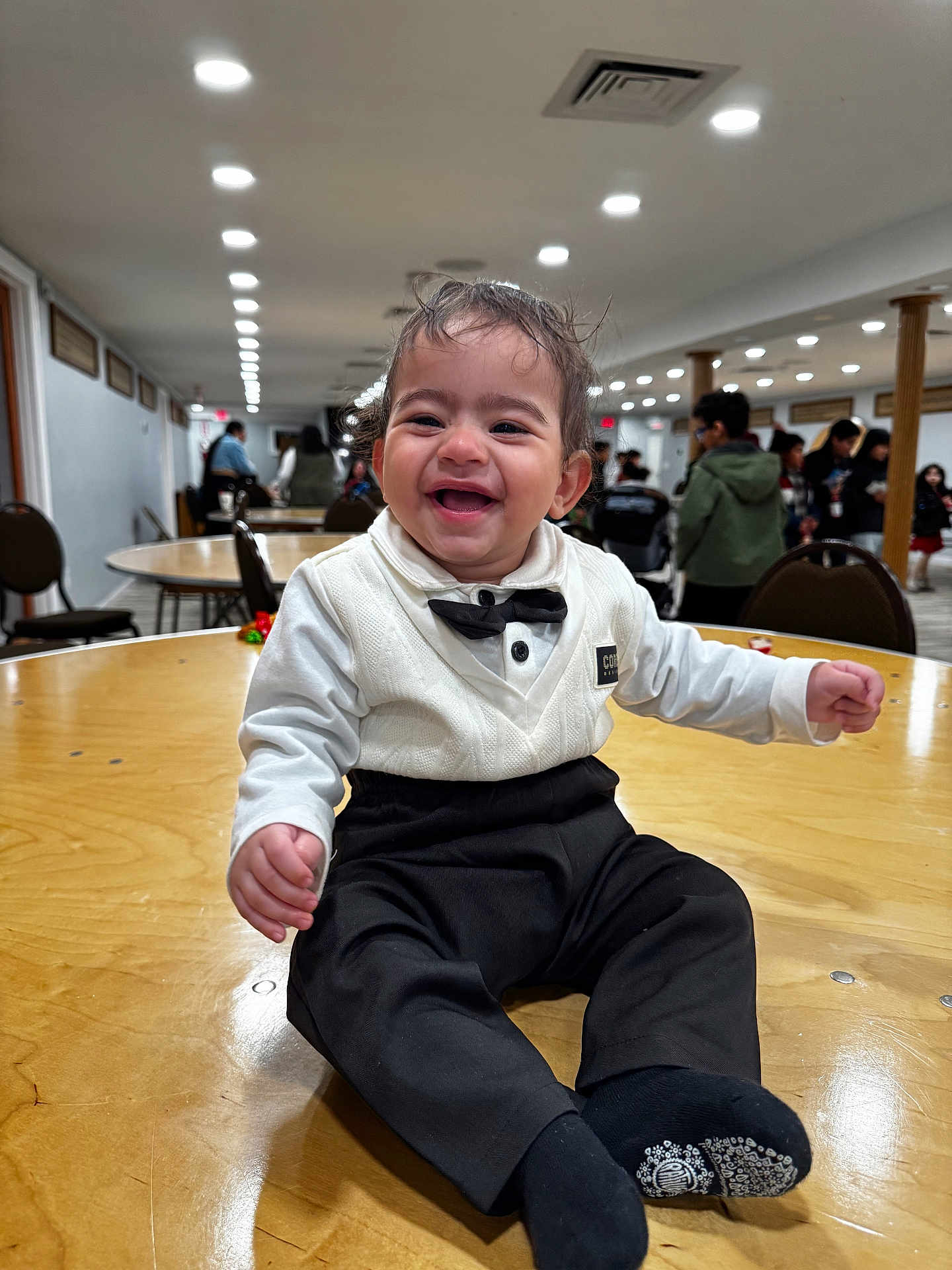 Augustine joined the competition — help win amazing prizes! baby, toddler, child, smiling, bow_tie, formal_attire, white_shirt, black_pants, socks, wooden_table, indoor, party, banquet_hall, people, chairs, ceiling_lights, happy, portrait, sitting, crowd
