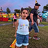child, girl, popcorn, fairground, outdoor, grass, blue_outfit, flowers, people, evening, casual_clothing, cap, shorts, sandals, trees, sky, festival, fun, family, event
