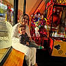 woman, toddler, child, arcade_game, bench, window, reflection, smile, casual_clothing, indoor, sunlight, colorful, playful, game_machine, happy, seated, family, joy, person, leisure