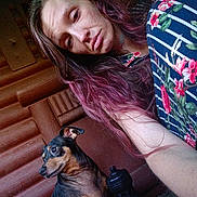 Lil Round Boy is registered to the contest to win money with this photo: woman, dog, pet, selfie, indoor, wooden_wall, log_cabin, casual, long_hair, purple_hair, striped_shirt, floral_pattern, small_dog, alert, portrait, companion, brown, black, relaxed, cozy