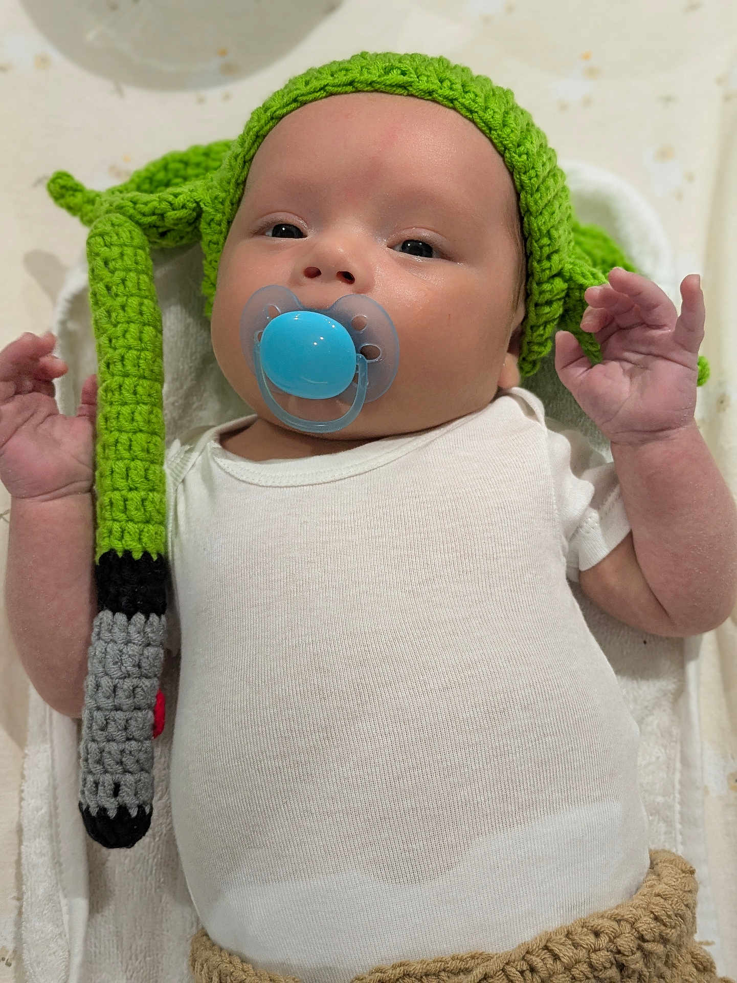 ézekiel a rejoint le concours — aidez-le/la à gagner de superbes lots ! baby, infant, pacifier, knitted_hat, yoda_hat, onesie, white_clothing, blanket, lying_down, hand, cute, child, soft_texture, closeup, indoors, newborn, portrait, calm, blue_pacifier, cozy