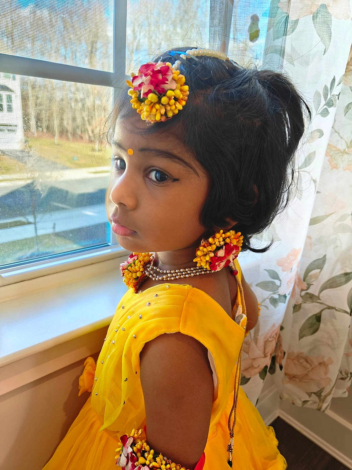 Thanmathi joined the competition — help win amazing prizes! child, girl, yellow_dress, floral_jewelry, window, natural_light, indoor, traditional_clothing, portrait, curtain, flower_accessories, young_child, face, looking_away, home, decor, black_hair, pensive, necklace, arm